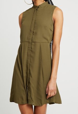 Vestido sin mangas verde oliva con cuello alto y parte frontal abotonada, llevado por una persona con trenza y grandes pendientes de aro.