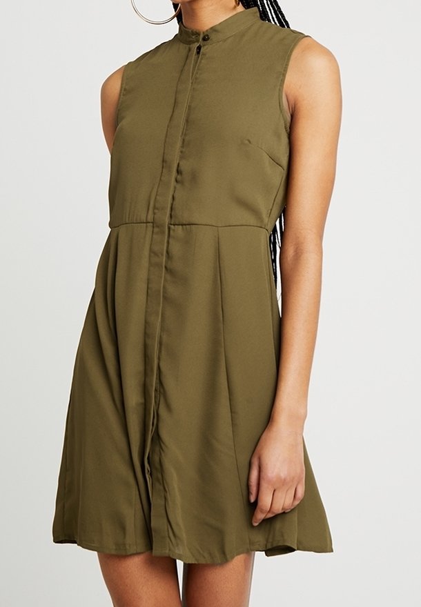 Robe sans manches vert olive avec col montant et devant boutonné, portée par une personne aux cheveux tressés et portant de grandes boucles d'oreilles anneaux.