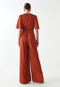 Mono de color óxido con mangas cortas abullonadas, cintura ajustada y pantalones de pierna ancha. Tejido suave y liso con cierre de cremallera en la parte posterior.