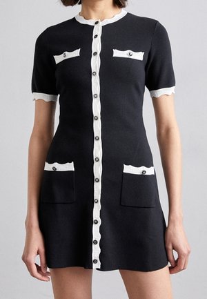 Schwarzes kurzärmeliges Kleid mit weißem, gewelltem Besatz, durchgeknöpft vorne und vier dekorativen Taschen mit Knöpfen, getragen von einer stehenden Person.