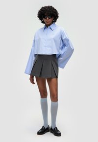 Lyseblå cropped skjorte med store ærmer, grå plisseret nederdel, lyseblå knæhøje strømper og sorte loafers, stylet med solbriller.
