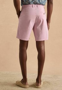 Helle rosa Shorts mit einer festen Textur, ausgestattet mit einer Gesäßtasche und einem kleinen Knopfdetail. Getragen mit einem gemusterten blauen Hemd.