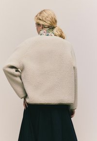 Kræmfarvet, struktureret fleecejakke med en afslappet pasform og raglanærmer, parret med en blomsterkrave og en mørk nederdel.