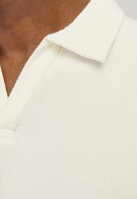 Camisa polo de canalé en beige claro con un cuello clásico y un cierre de botones. La tela parece suave y texturizada con líneas sutiles.
