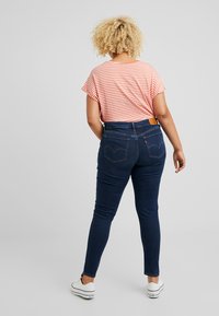 Persona con cabello rizado rubio, vistiendo una camiseta a rayas rojas y blancas, jeans ajustados azul oscuro y zapatillas blancas, de pie de espaldas.