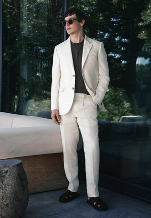 Mand iført cremefarvet jakkesæt, mørk skjorte, solbriller og sorte sandaler står indendørs nær en beige sofa og et glasvindue, der reflekterer træer.