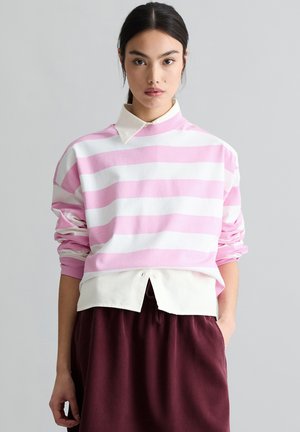 Młoda kobieta o ciemnych włosach, ubrana w różowo-biały sweter w paski na białej koszuli z kołnierzykiem oraz bordowe spodnie, stojąca na tle szarego tła.