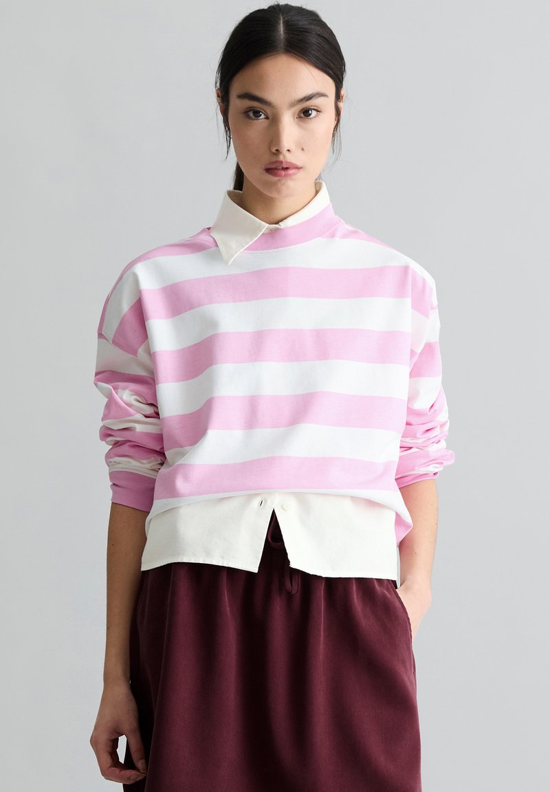 Młoda kobieta o ciemnych włosach, ubrana w różowo-biały sweter w paski na białej koszuli z kołnierzykiem oraz bordowe spodnie, stojąca na tle szarego tła.