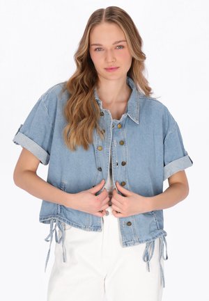 Jonge vrouw met een cropped lichtblauwe spijkerjack met opgerolde mouwen en witte broek, poserend tegen een witte achtergrond.