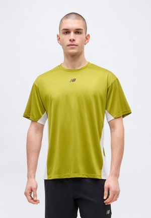 Noor mees, kes kannab rohelist New Balance lühikeste varrukatega spordisärki ja musta lühikesi pükse, seisab ühevärvilise heleda tausta ees.