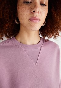 Lilla genser med rund hals, ribbet krage og en subtil V-formet design detalj på fronten. Mykt stoff med en glatt tekstur.