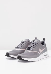 Pár šedých a růžových tenisek Nike Air Max Thea s bílou podrážkou na bílém povrchu a jednoduchém pozadí.