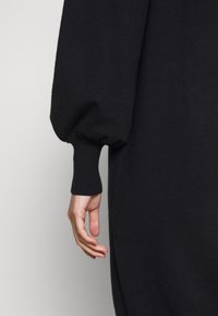 Robe en maille noire avec une manche bouffante ample et un poignet ajusté. Design sans couture avec une texture lisse et un drapé subtil. Main reposant sur le côté.