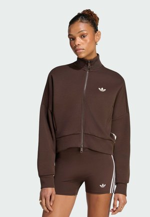 Frau mit lockigem Haar zu einem Dutt gebunden trägt eine braune Jacke mit Reißverschluss und passende Shorts mit weißen Adidas-Logos und Streifen.