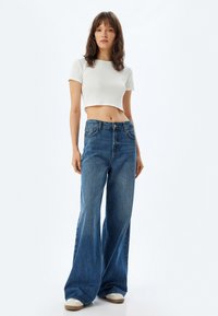 Hoge taille wijde jeans in medium blauwe denim, met een ritssluiting, vijf zakken en een relaxed fit; gecombineerd met een wit cropped top.