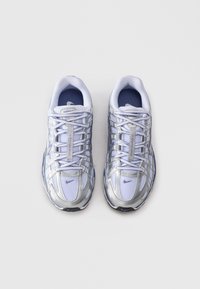 Sneakers atletici bianchi e argentati con tessuto a rete e lacci incrociati, mostrati dall'alto su uno sfondo chiaro e semplice.