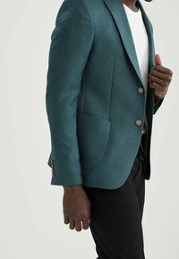Blazer testurizzato color teal con colletto a punta, due bottoni e una tasca sul petto, abbinato a una t-shirt bianca e pantaloni neri.