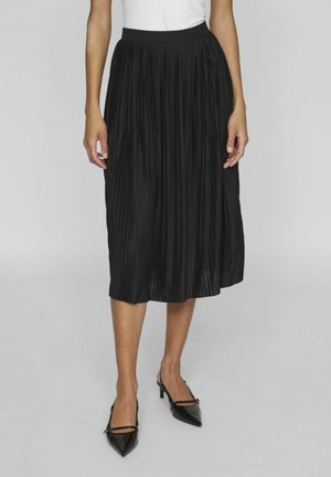 Kobieta w czarnej plisowanej spódnicy midi z wysokim stanem, w czarnych szpilkach na pasku z zakrytymi palcami, stojąca na jednolitym tle.