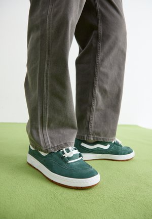 Green suede sneakers with white accents, rubber sole, and white laces, paired with gray wide-leg pants, on a light green surface.