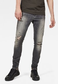 Vaqueros skinny fit en denim gris desgastado. Presentan detalles desgastados y múltiples roturas. Combinados con zapatillas negras y una camiseta verde.