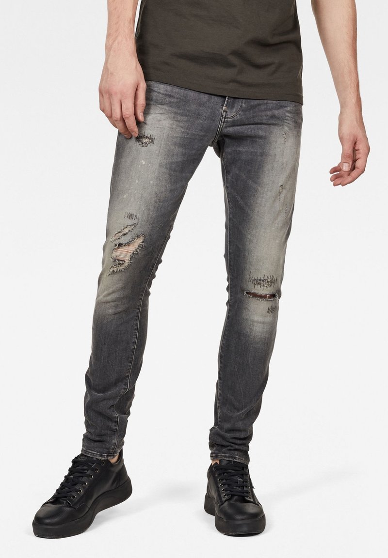 Vaqueros skinny fit en denim gris desgastado. Presentan detalles desgastados y múltiples roturas. Combinados con zapatillas negras y una camiseta verde.