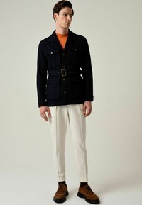 Marineblauw jasje met ceintuur en vier voorzakken, gecombineerd met witte taps toelopende broek en bruine suède schoenen. Oranje overhemd eronder.