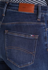 Mörkblå denimjeans med en brun läderetikett med "HILFIGER DENIM", kontrasterande sömmar och en liten dekorativ flagga på bakfickan.