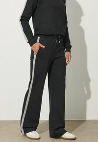 Schwarze Jogginghosen mit grauen Seitenstreifen, aus weichem Stoff gefertigt. Sie verfügen über einen elastischen Bund, einen Kordelzug und Seitentaschen.