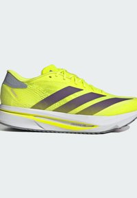 Des chaussures de sport néon jaunes avec des accents gris, présentant une tige en mesh, trois rayures violettes foncées, et une semelle légère.
