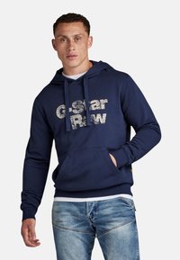 Navy blue hoodie with a front kangaroo pocket, featuring a distressed white "G-Star Raw" logo. Made of soft, textured fabric suitable for casual wear.