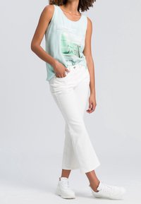 Mujer vestida con una blusa sin mangas de color azul claro, jeans acampanados cortos blancos y zapatillas blancas, de pie con una mano en el bolsillo.
