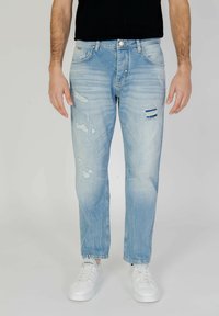 Jeans de mezclilla azul claro con un corte relajado, que presentan desgastes, arrugas y parches rasgados. Combinados con zapatillas blancas.