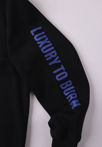 Μαύρο φούτερ με μακριά μανίκια, ριμπ στο τελείωμα και έντονη μπλε επιγραφή "LUXURY TO BURN" κατά μήκος του αριστερού μανικιού.