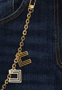 Collana d'oro con due charm geometrici: una "C" aperta e un quadrato solido, attaccata a un tessuto di denim blu scuro con cuciture gialle.