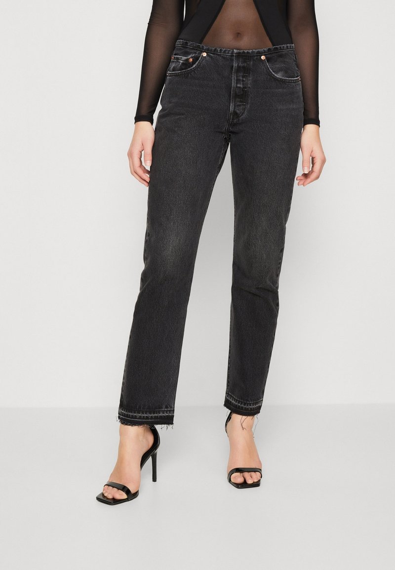 Femme portant un jean noir taille haute à ourlets bruts et des sandales à talons hauts noires à bout ouvert, debout devant un fond uni.