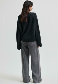 Sieviete ar gariem brūniem matiem, tērpta brīvā melnā džemperī un platās pelēkās džinsās, stāv vērsta prom pret vienkāršu gaišu fona.