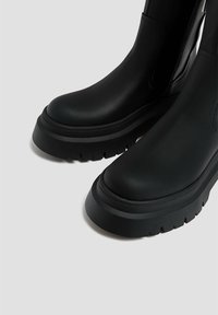 Botas negras hasta el tobillo hechas de cuero suave con una suela de plataforma gruesa, con punta redondeada y cierre de cremallera lateral. Detalle de suela texturizada.