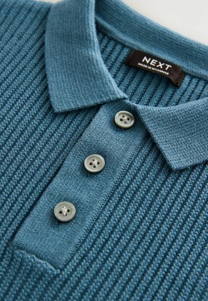 Polo-shirt bleu texturé avec un motif cannelé, trois boutons gris et un col. L'étiquette indique "NEXT, fabriqué au Myanmar."