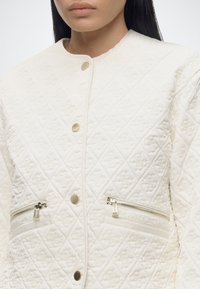 Femme portant une veste matelassée blanche avec des motifs en losange, des boutons-pression dorés et des poches avant zippées, montrée des épaules jusqu’au milieu du torse.