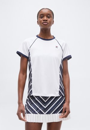 Ženska stoji oblečena v belo in mornarsko modro športno opravo z vzorci v obliki cik-cak črt in s plisirano krilo na enobarvnem svetlem ozadju.