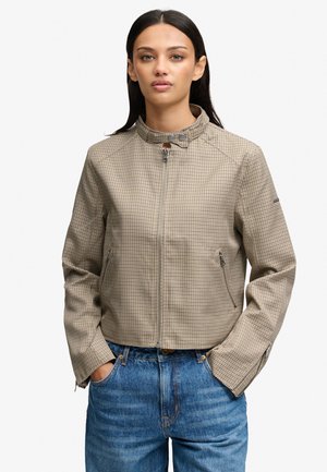 Kvinde iført en beige ternet lynlåstrøje med trykknaplukning og blå jeans, stående med hænderne i lommerne og neutralt ansigtsudtryk.