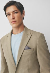 Béžový kordový blazer s klopami a náprsní kapsou, obsahujúci tmavo vzorovanú vreckovku. Pod tým svetlomodré tričko.
