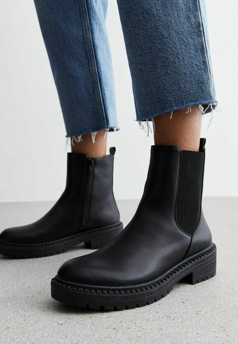 Bottines en cuir noir avec semelles épaisses à crampons portées avec un jean bleu à ourlet effiloché sur un sol gris.