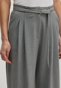 Grijze geplooide broek van gestructureerde stof, met een bretel tailleband met een zilveren ring en zijzakken.