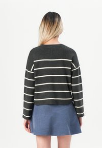 Frau mit langen blonden Ombre-Haar, die einen dunkelgrauen Pullover mit weißen horizontalen Streifen und einen blauen Rock trägt, von hinten gezeigt.