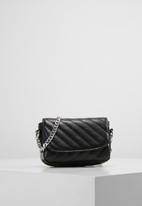 Borsa a tracolla in pelle trapuntata nera con tracolla in catena argentata, posata su un pedestal bianco su uno sfondo grigio semplice.