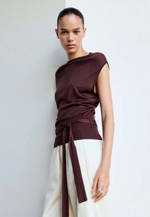 Jeune personne aux cheveux plaqués en arrière portant un haut sans manches bordeaux avec une longue ceinture et un pantalon large de couleur claire, debout contre un mur uni.