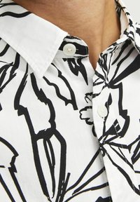 Chemise blanche à boutons, ornée d'un motif floral noir, avec un col pointu et un tissu texturé. Deux boutons visibles au niveau du col.