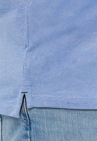 Lichtblauw shirt van een gestructureerde stof met zijsplitten. Afgewerkte randen en een klein zwart stiksel aan de onderzijde aan de linkerkant.