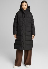 Manteau en duvet noir avec capuche, présentant un design matelassé, une fermeture éclair avant et des poches latérales. Porté sur un pantalon large marron et des chaussures blanches.
